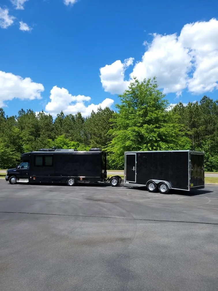 A black Class C motorhome towing a black enclosed car hauler in a parking lot, with a Trailer Toad between them.