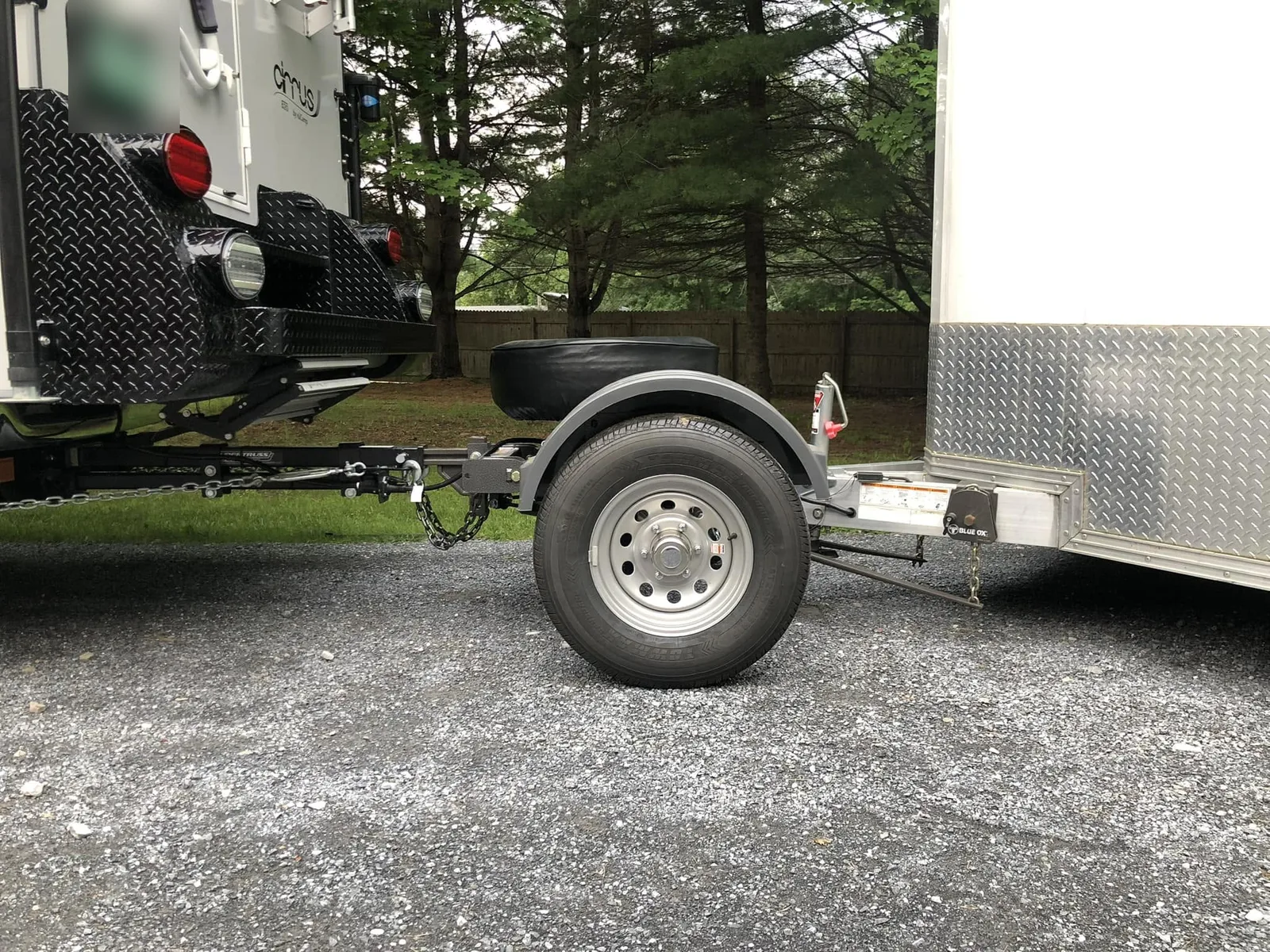 Close-up of a Trailer Toad hitched between the back of a truck camper and the front of a white enclosed trailer. The weight distribution hitch’s shank runs from the camper’s receiver to the front of the Toad, the Toad sits on one wheel under a fender, and the trailer couples to the weight distribution hitch at the rear of the Toad.