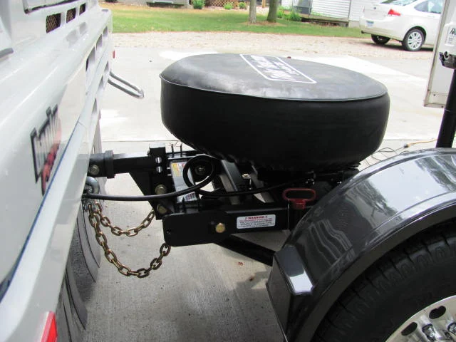 Front of a Trailer Toad hitched to a motorhome: the weight distribution hitch shank sits in the receiver with safety chains attached, the spare tire rides on top, and a red-handled reverse pin is visible on the side.
